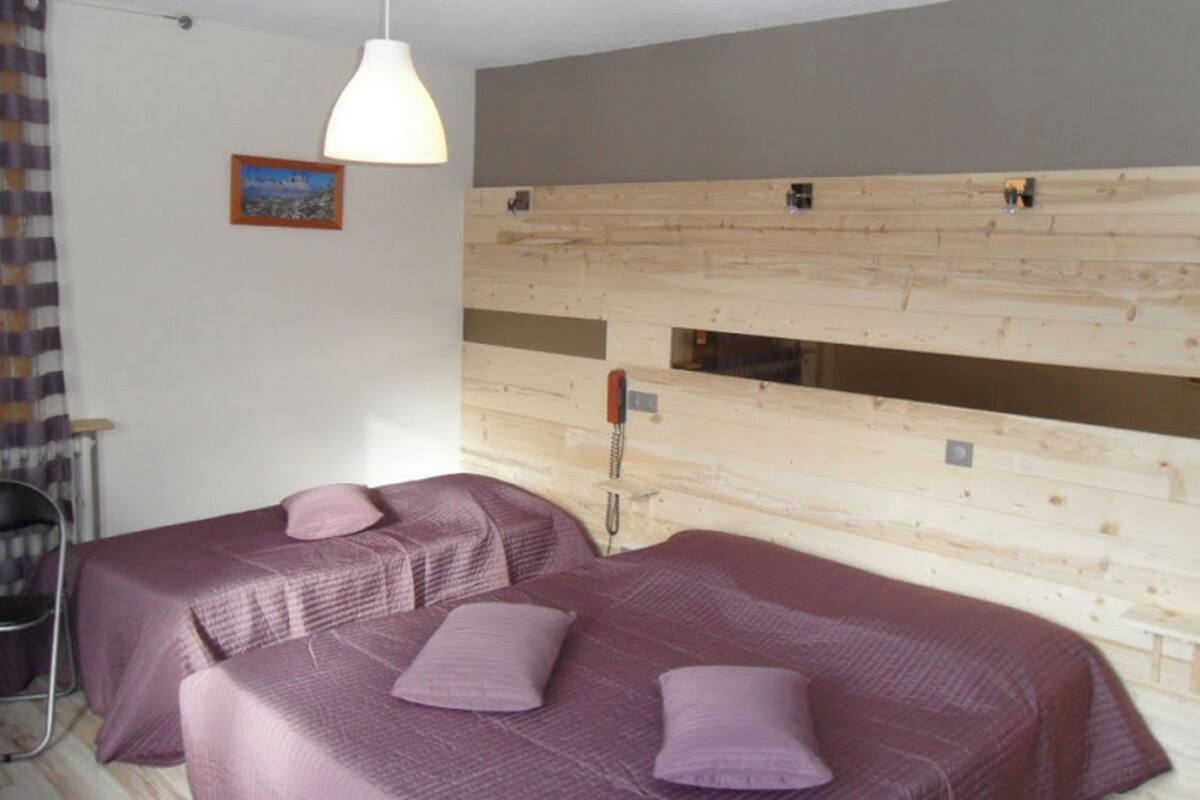 A room with two beds, each with a purple bedspread and pillows, a wooden headboard, a hanging light fixture, and a small window.