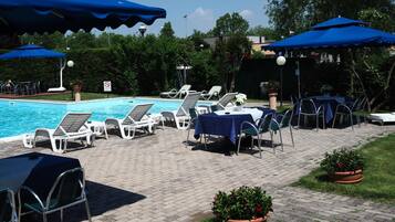 Zona junto a la piscina con sillas de descanso y sombrillas, piscina y plantas en macetas.
