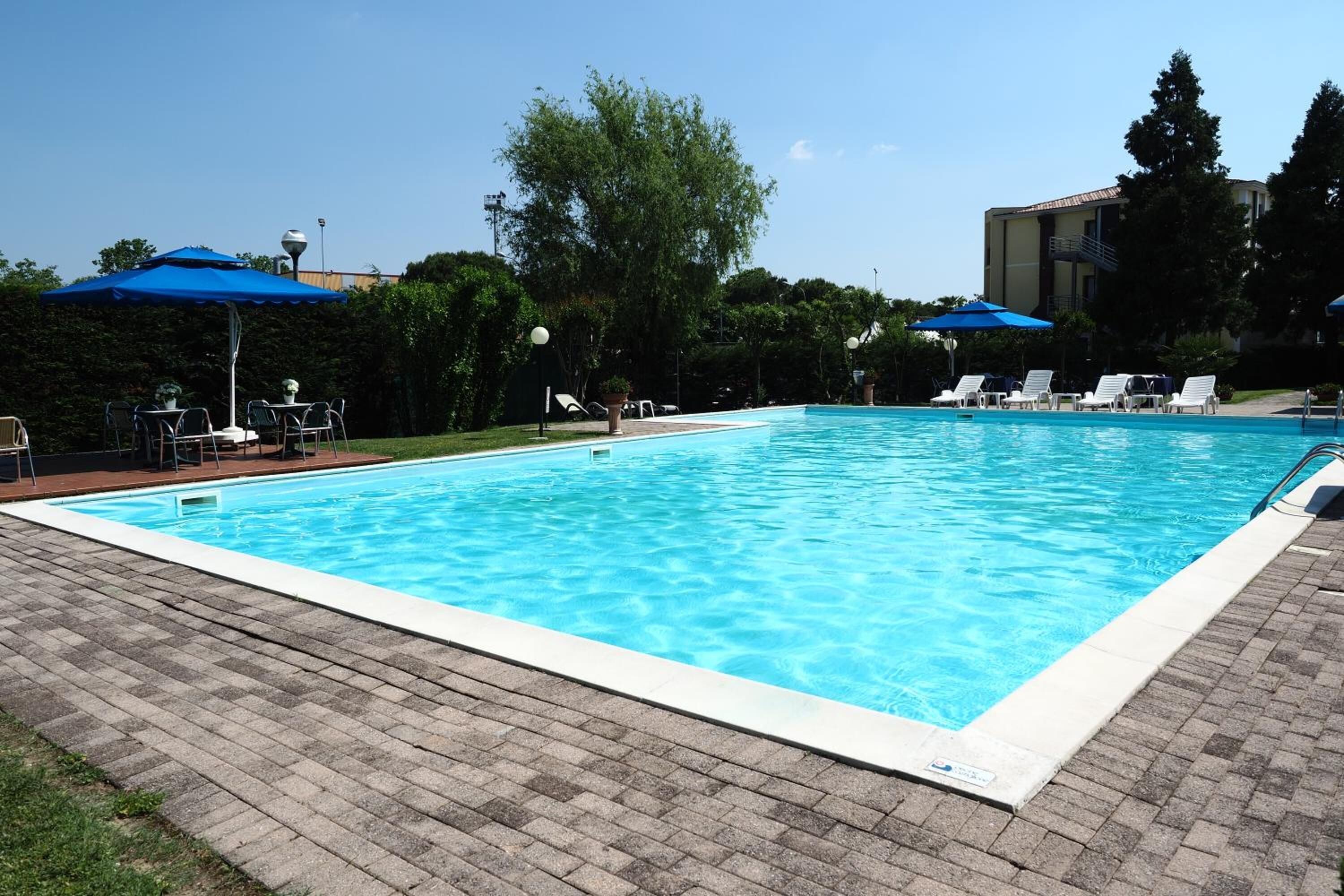 En rektangulär swimmingpool med klart, blått vatten, omgiven av en stenlagd terrass. Längs poolkanten finns solstolar och parasoller.