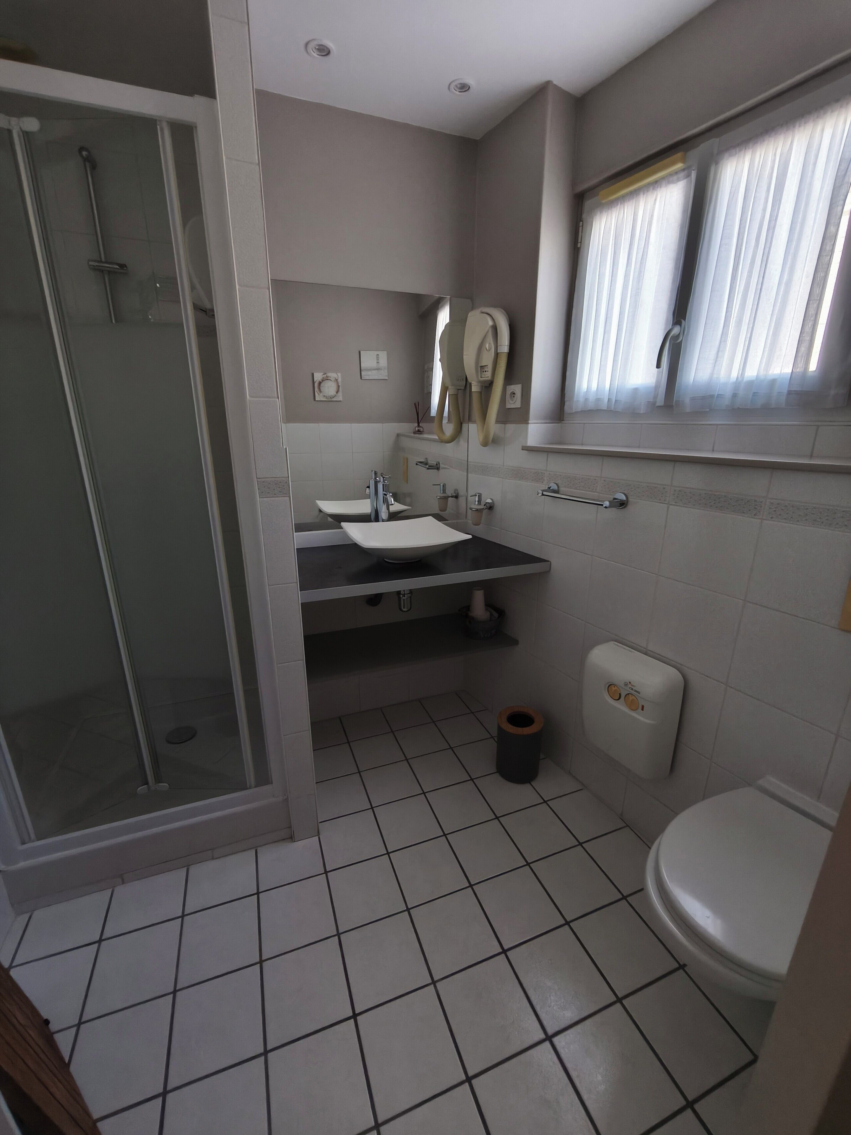 Bagno con doccia, due lavabi, un water e una finestra con tende.