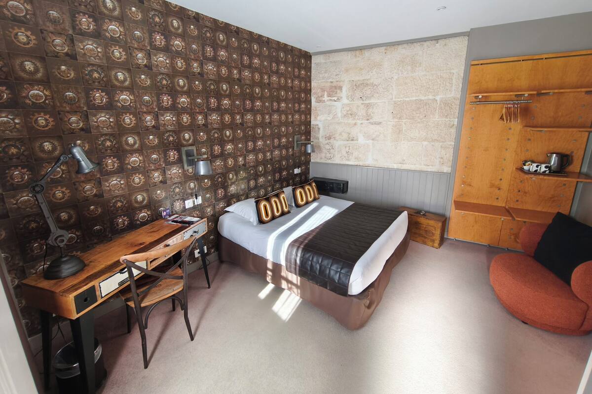 Un dormitorio con pared estampada, una cama con cabecera original, un escritorio con lámpara, una silla y un armario.