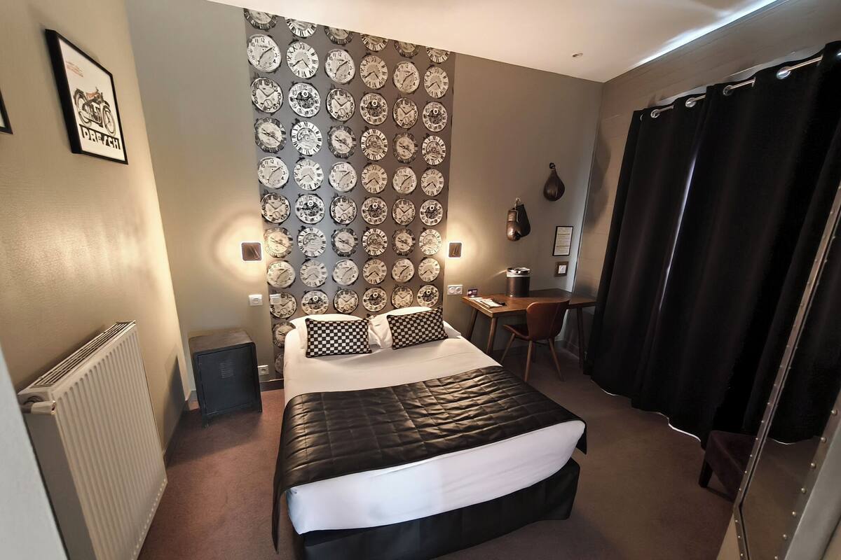 Habitación de hotel con cama, escritorio, silla, radiador y una pared con relojes.