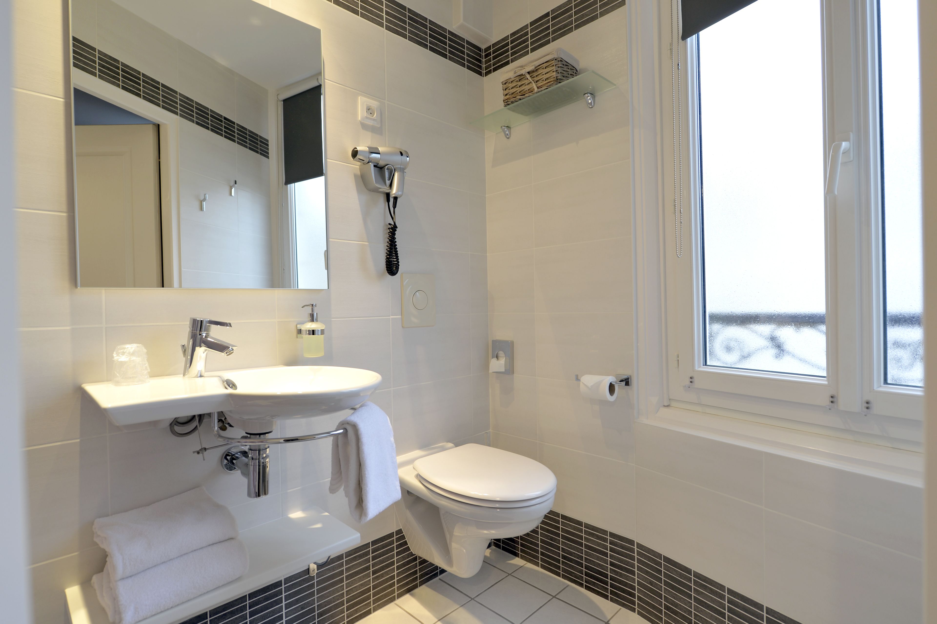 Un bagno moderno con lavandino bianco, specchio e water. Presente un portasciugamani con asciugamani piegati, un asciugacapelli montato a parete e una finestra con vista esterna.