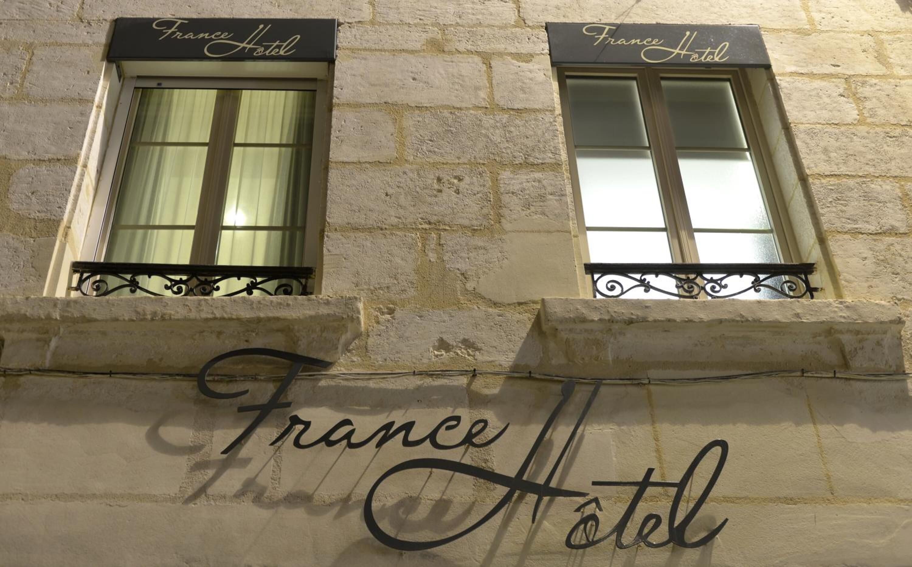 La façade d’un immeuble présente deux fenêtres portant l’inscription « France Hôtel », chacune étant ornée de balustrades en fer forgé.