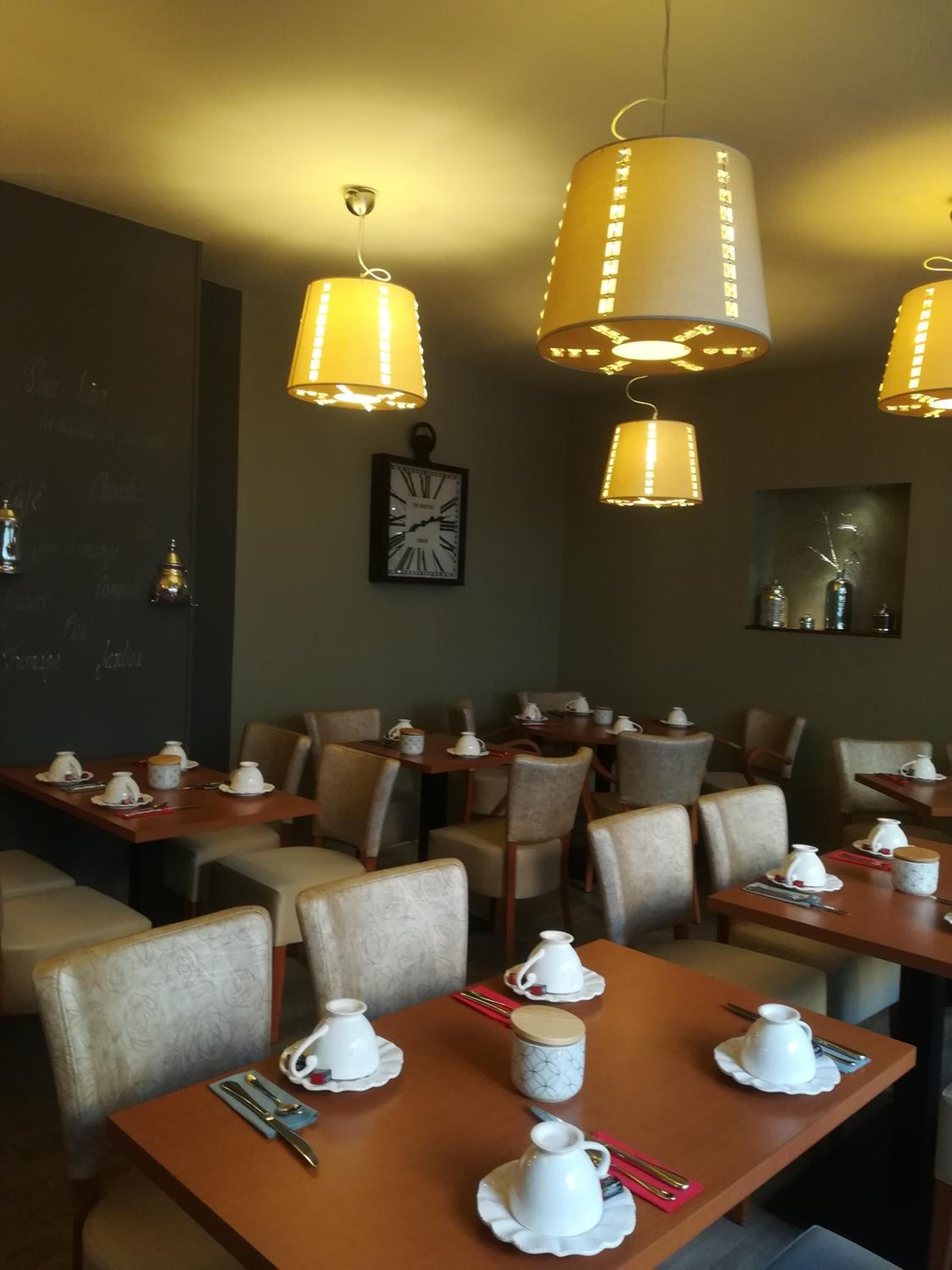 Un comedor con mesas preparadas para el desayuno, con teteras y tazas blancas, y lámparas colgantes arriba.