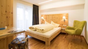A wooden bed with a headboard, two bedside lamps, a green armchair, and a wooden nightstand with a lamp. - Pension Hilpold (Lana)