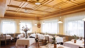 A restaurant with multiple tables and chairs, wooden ceiling, and large windows with sheer curtains. - Pension Hilpold (Lana)