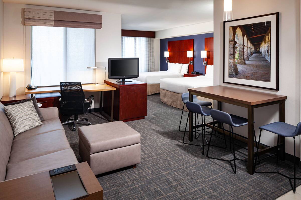 Habitación de hotel con sofá, escritorio, televisor, cama y zona de comedor con sillas.