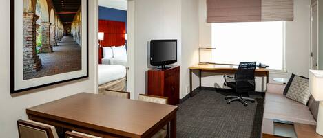 Habitación de hotel con cama, escritorio con computadora, televisor, mesa de comedor con sillas y un gran ventanal con vista a un edificio.