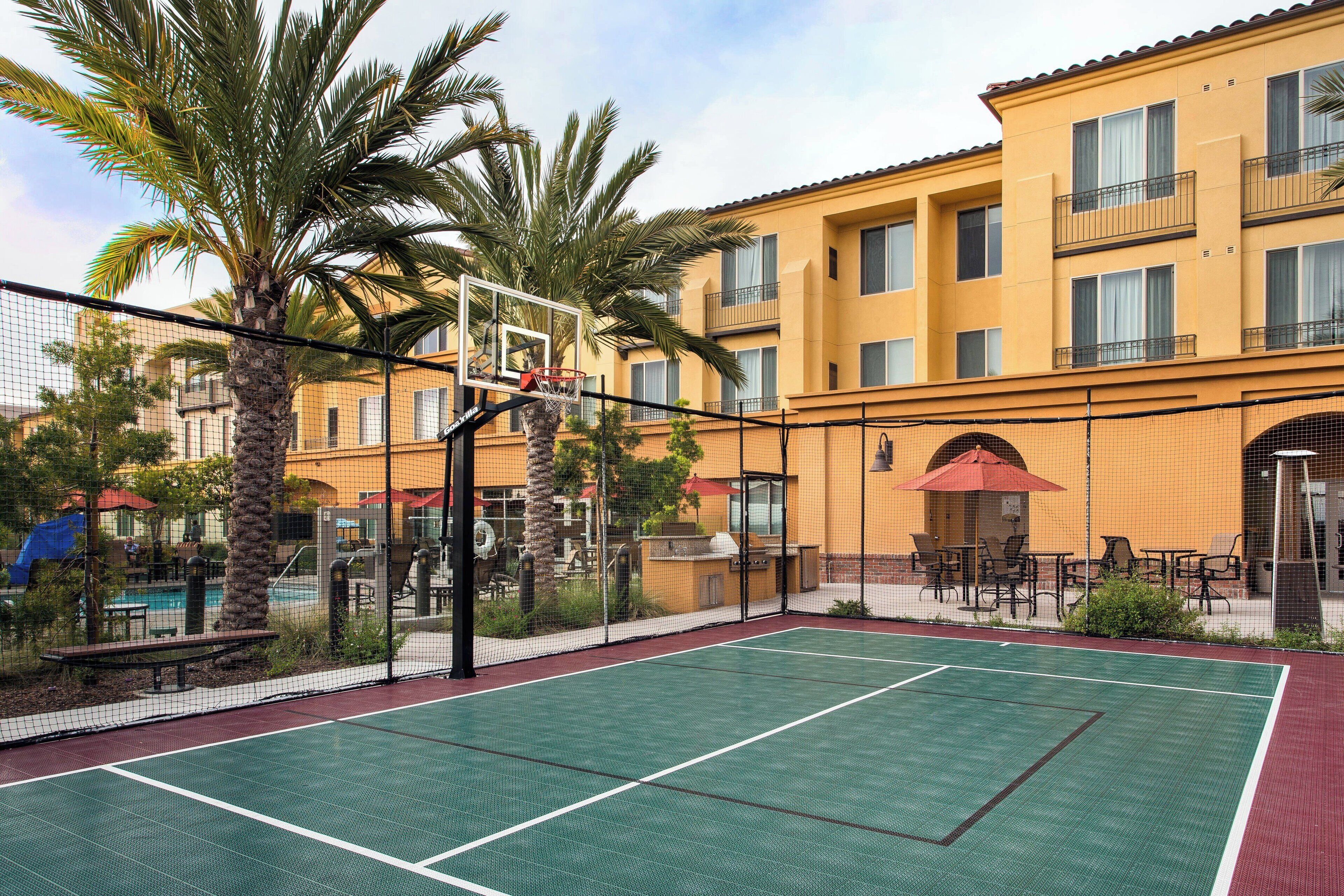 An outdoor tennis court with a basketball hoop, surrounded by palm trees and a pool area.