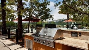 An outdoor kitchen area with a stainless steel grill, seating under umbrellas, and a view of the surrounding landscape.