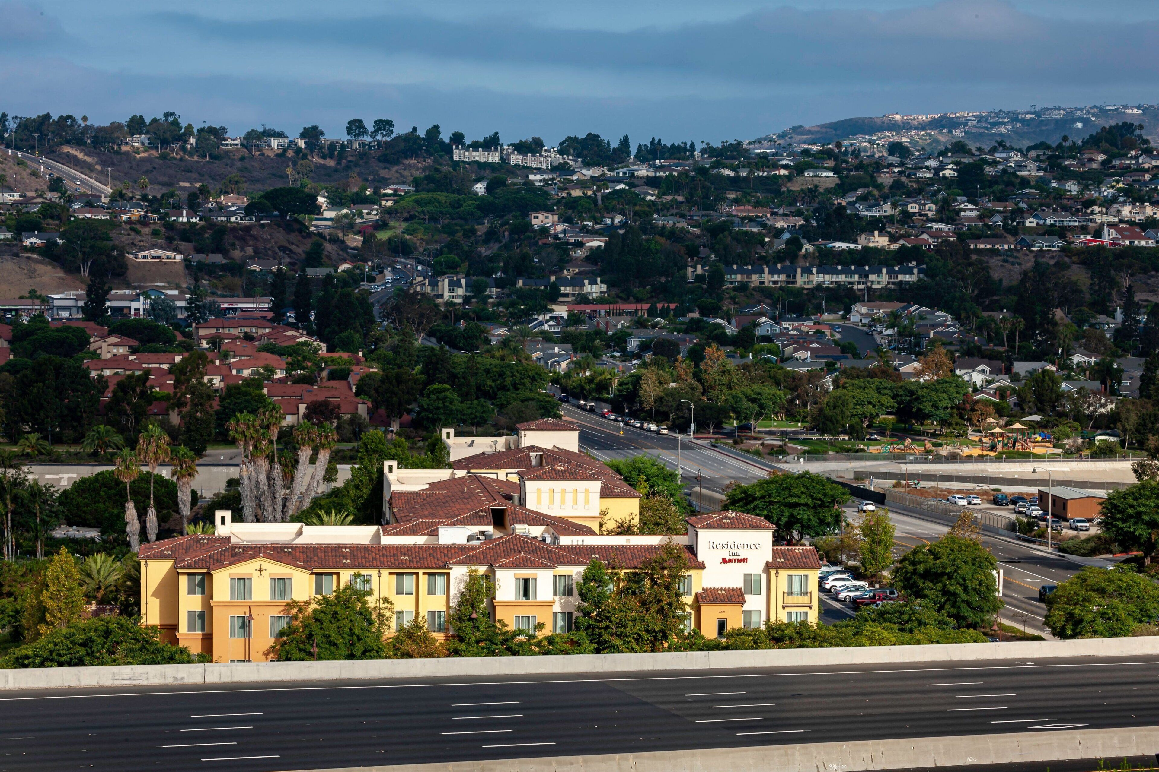 A residential area with a hotel named 'Residence Marriott' featuring a modern design, a parking lot, and a road leading to a hilltop area.