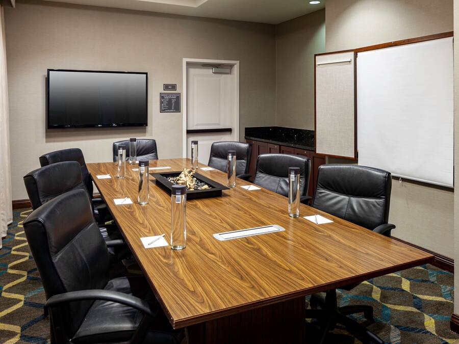 A conference room with a long wooden table, black leather chairs, a flat-screen TV, a whiteboard, and a patterned carpet.