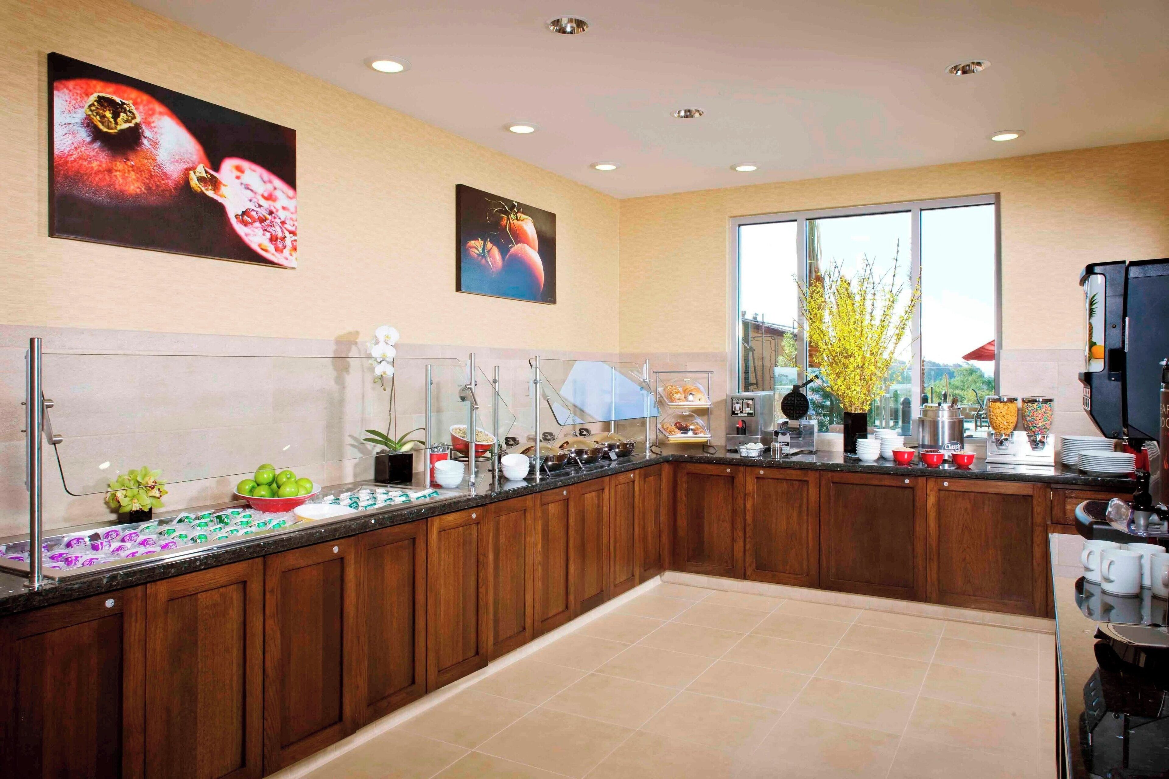 A modern kitchen with wooden cabinets, a glass counter, and various appliances. There are two large windows providing natural light and a view of the outdoors.