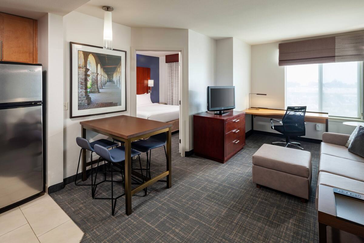 Habitación de hotel compacta con kitchenette, una cama, un escritorio con silla, televisión y un ventanal con vistas a la ciudad.