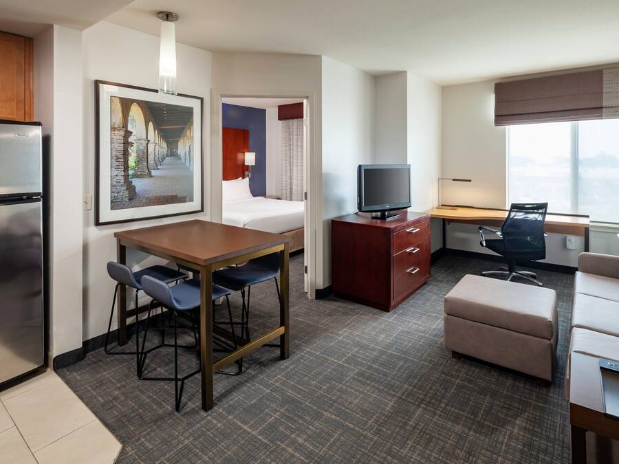 Une chambre d’hôtel compacte comprenant un coin cuisine, un lit, un bureau avec une chaise, une télévision et une grande fenêtre donnant sur la ville.