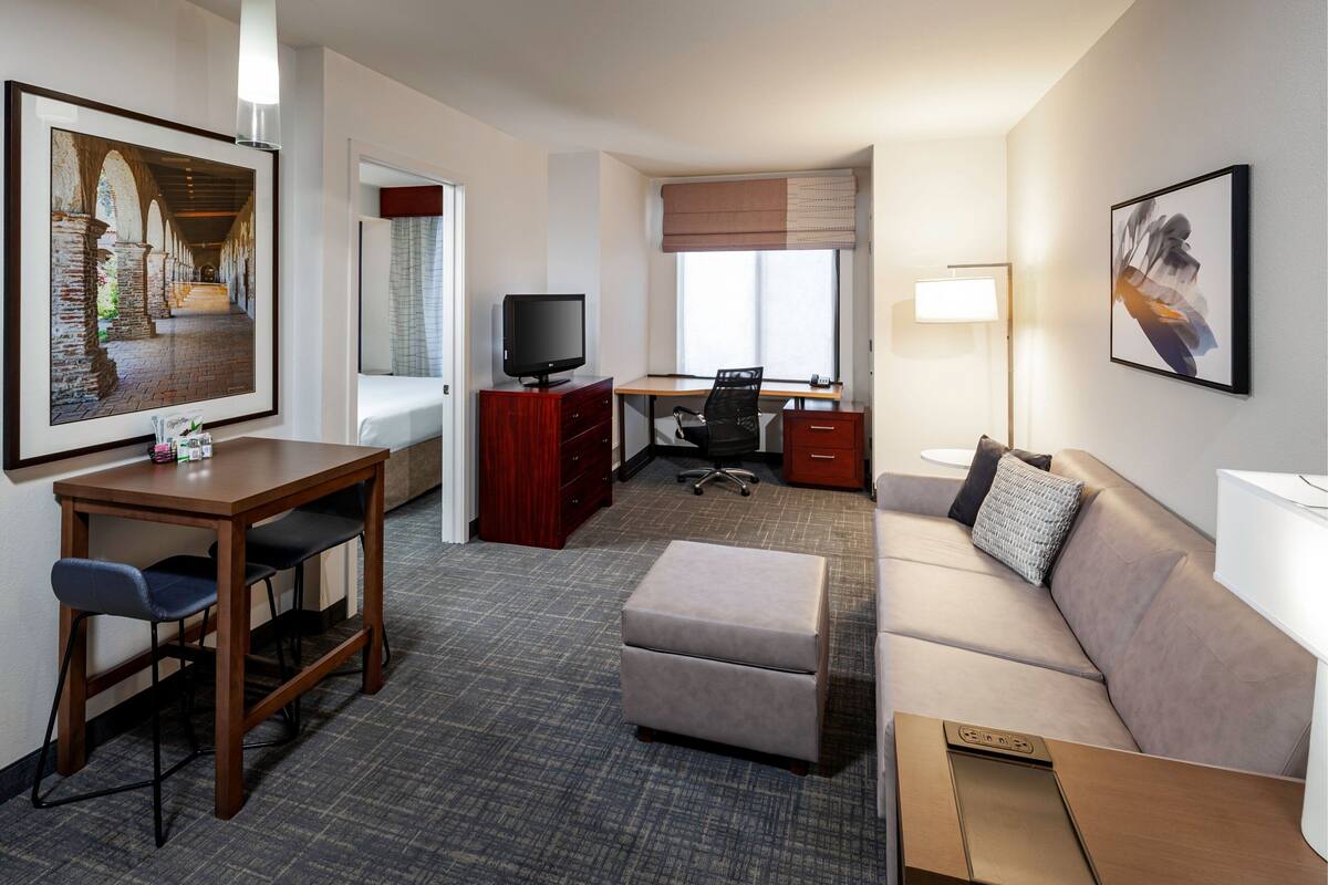 Habitación de hotel con escritorio, silla, televisor, sofá, otoman y obras de arte en las paredes.