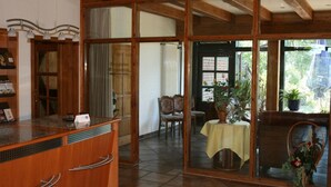 Un área de recepción con un mostrador de madera, una recepción y un separador de vidrio. Se observa una mesa con mantel amarillo y sillas al fondo.