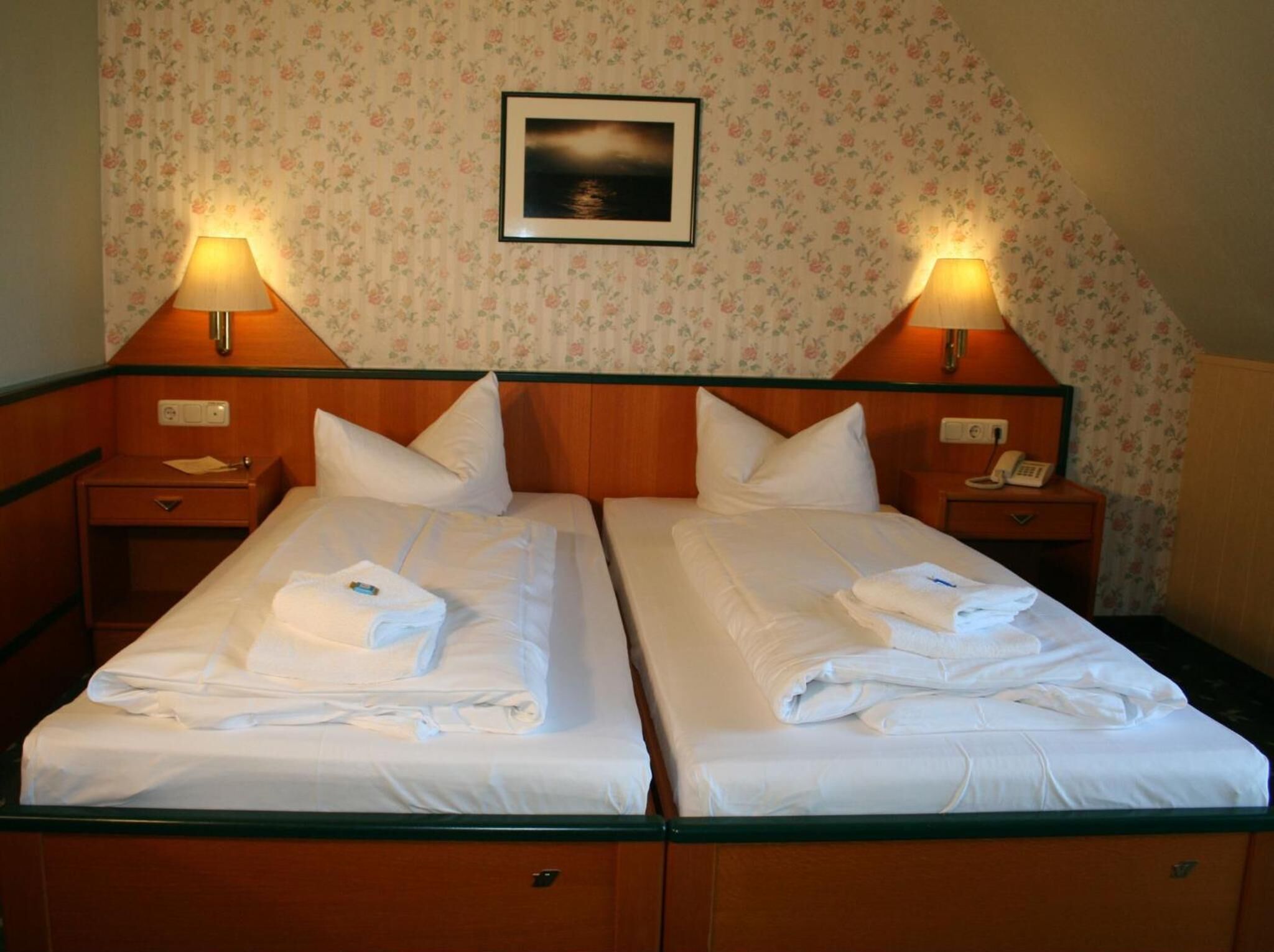 A hotel room with two beds, each with a pillow and a folded towel. There are two bedside lamps, a telephone on the right side, and a framed picture on the wall above the beds.