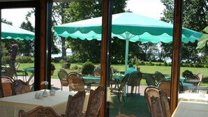Un área de comedor al aire libre con mesas y sillas, cubierta por grandes sombrillas verdes, y vista a un jardín bien cuidado.