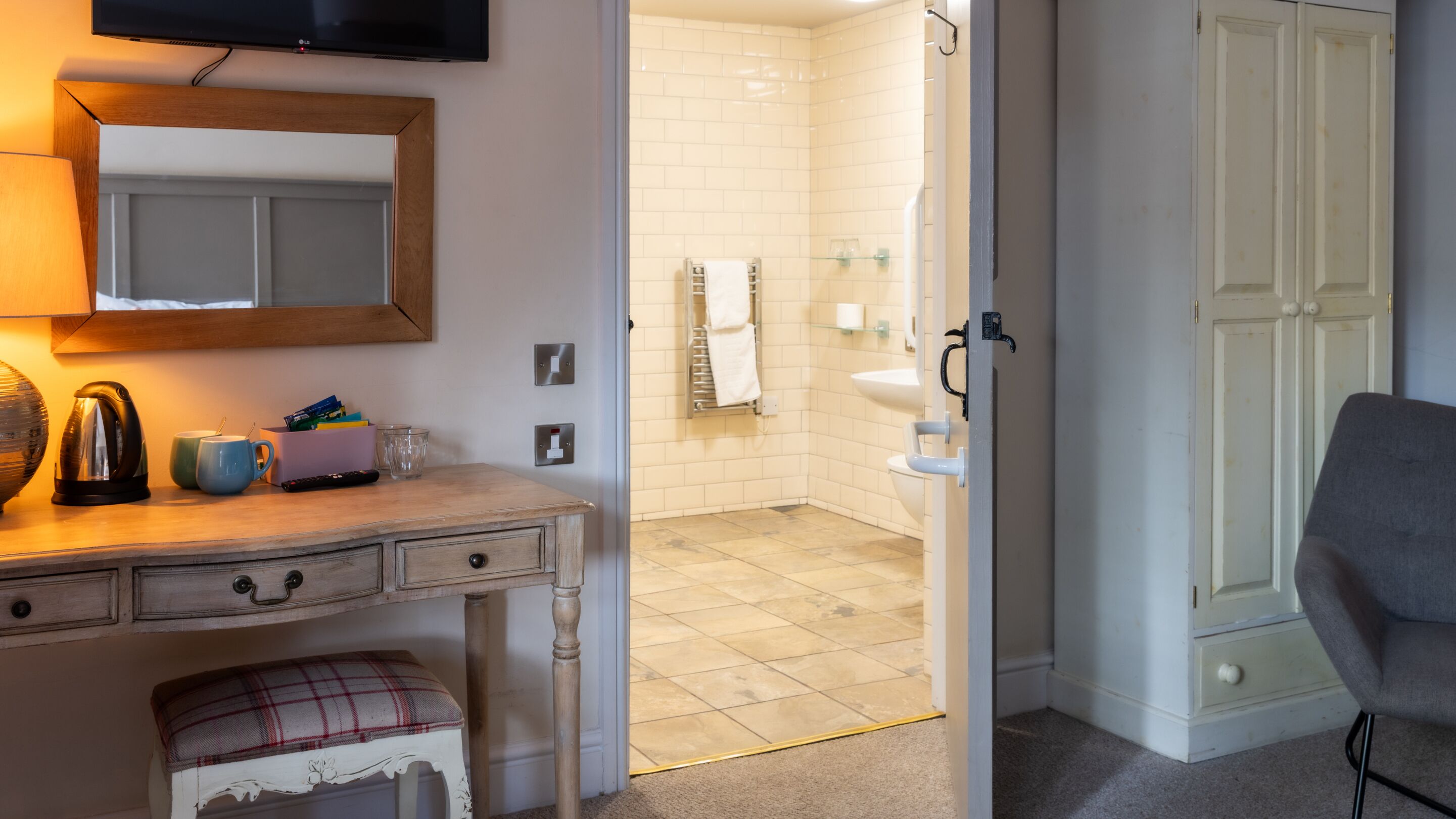 A hotel room with a wooden desk, a television, a mirror, a plaid bench, and a view into the bathroom with a sink and towel rack.
