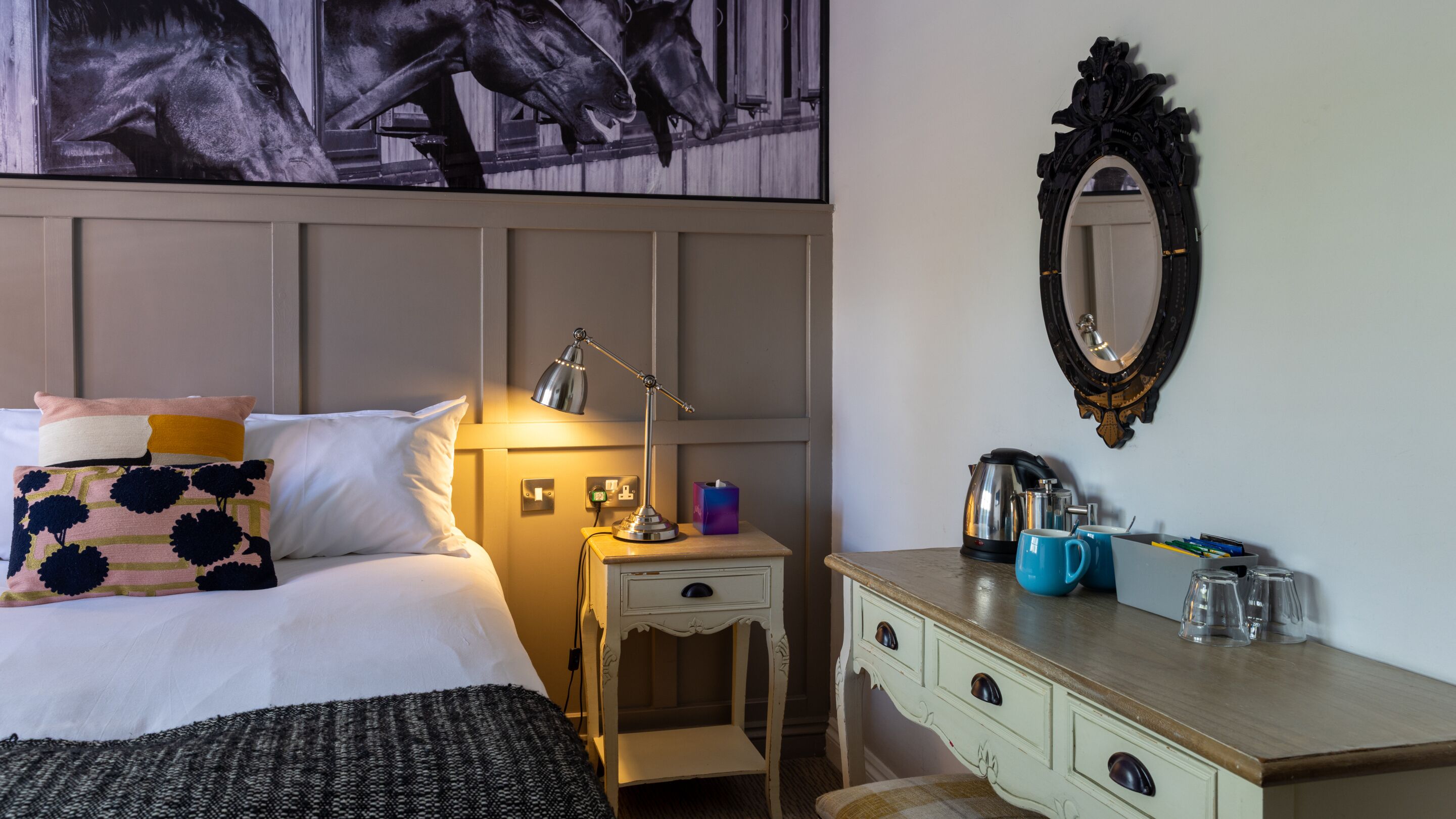Habitación de hotel con cama, mesita de noche, lámpara, espejo, cómoda y un cuadro de caballos.