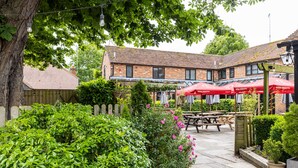 A courtyard with a brick building, wooden fence, and outdoor seating area with red umbrellas. - The Crown & Horns (Newbury)