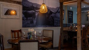 A dining area with a table set for two, a hanging light fixture, and a large framed picture of a bull. - The Crown & Horns (Newbury)