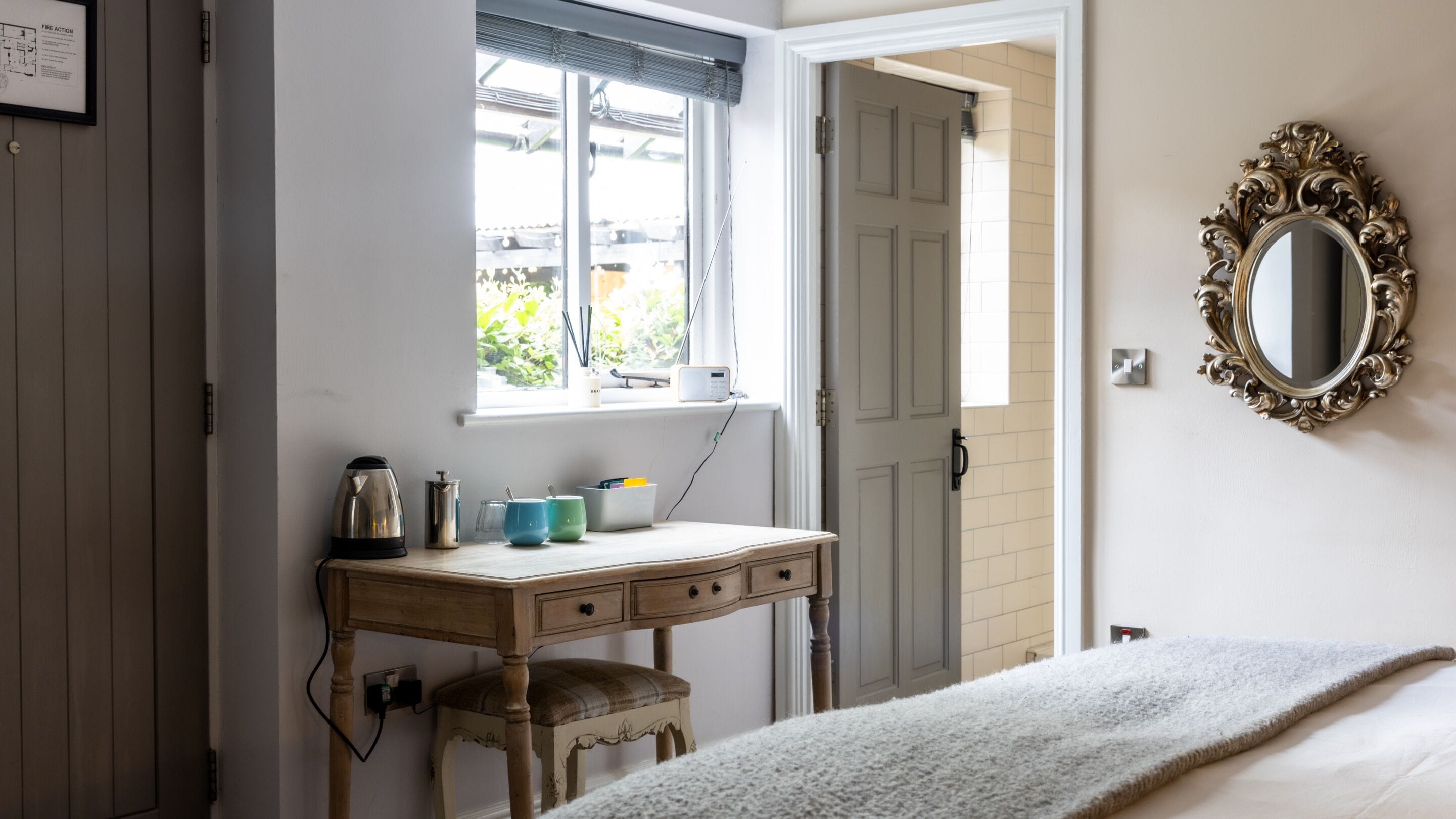 A bedroom with a wooden desk, a bed with a white blanket, a mirror with an ornate frame, and a window with blinds.