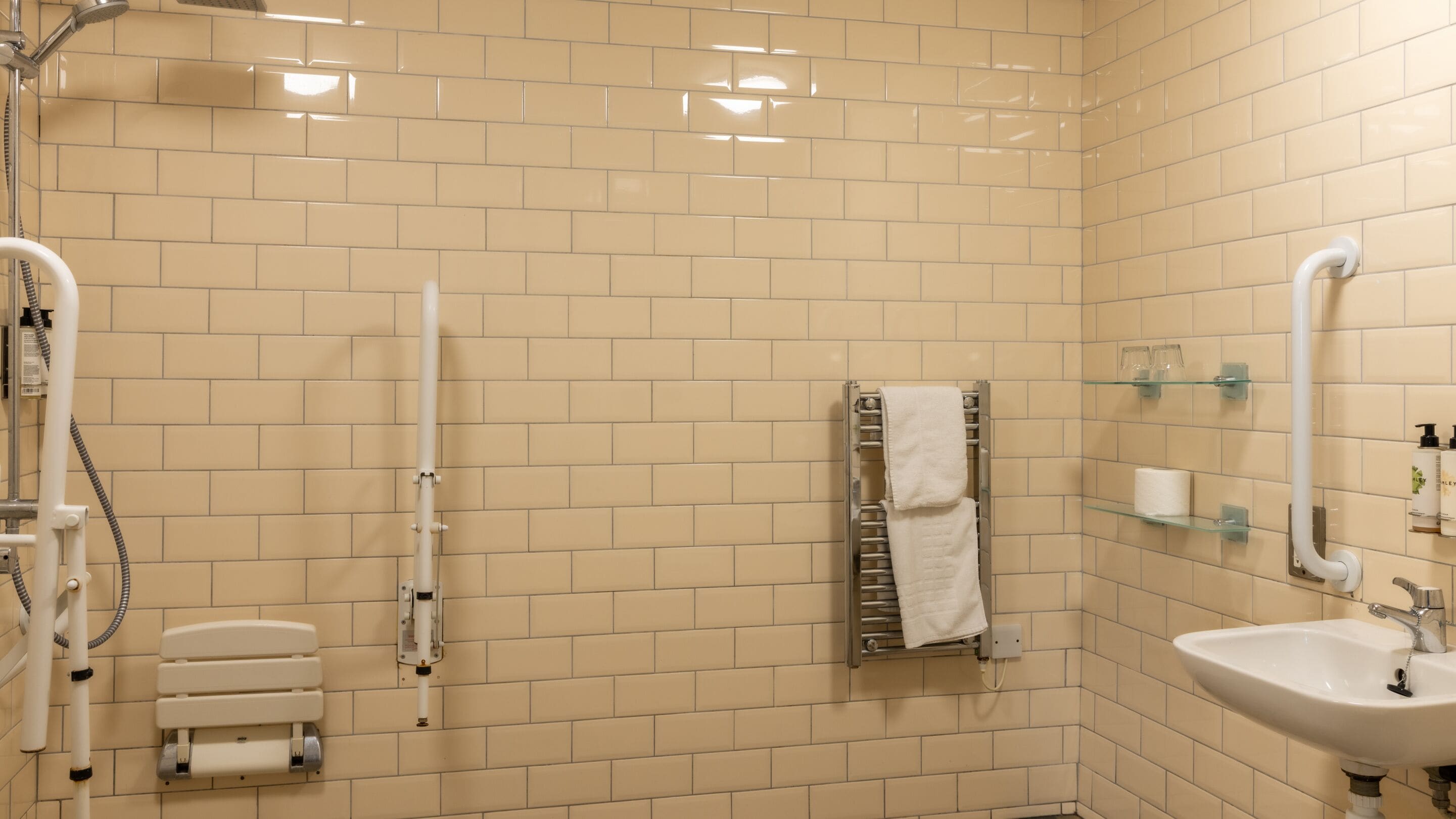 Un baño con paredes de baldosas color beige, un lavamanos blanco, un estante de vidrio con artículos de aseo, un perchero y una ducha con cabezal de ducha manual.