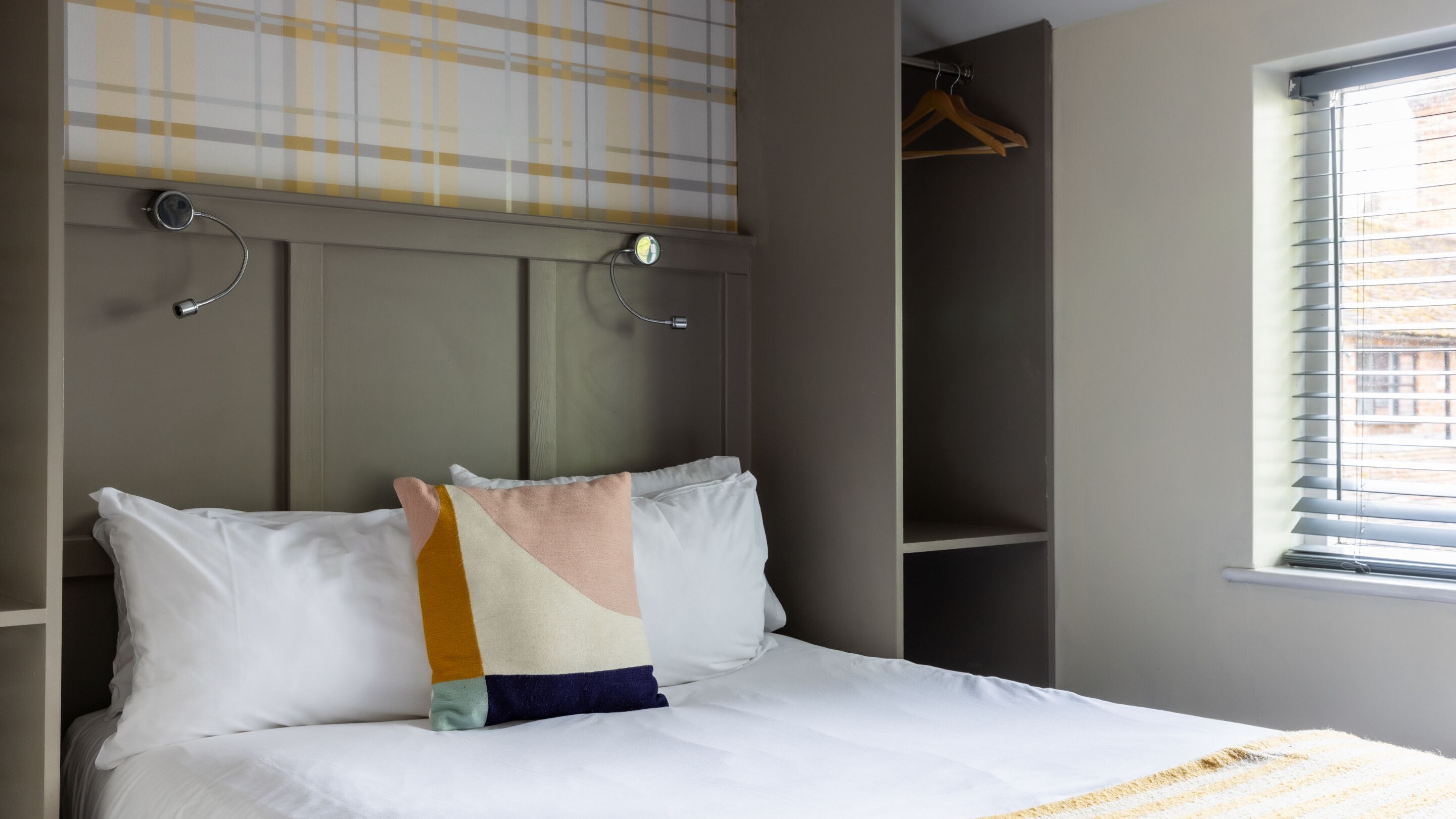 A bed with white bedding and a multicolored pillow, a bedside table, a hanging light, a wardrobe, and a window with blinds.