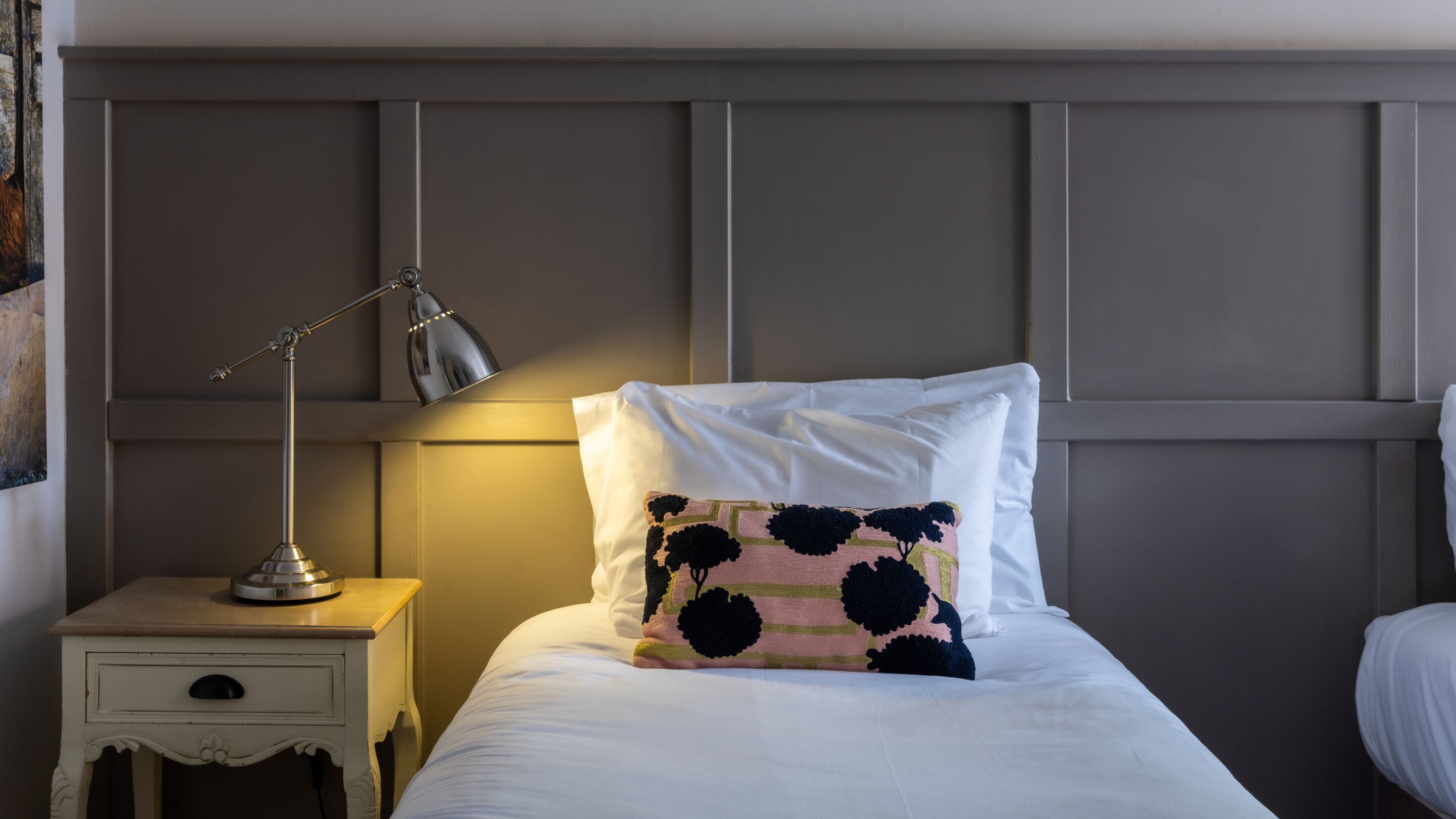 A neatly made bed with a white pillow and a decorative pillow with black and pink floral patterns. There is a bedside lamp on a small white nightstand with a drawer. The room has a simple, clean design with a grey headboard.