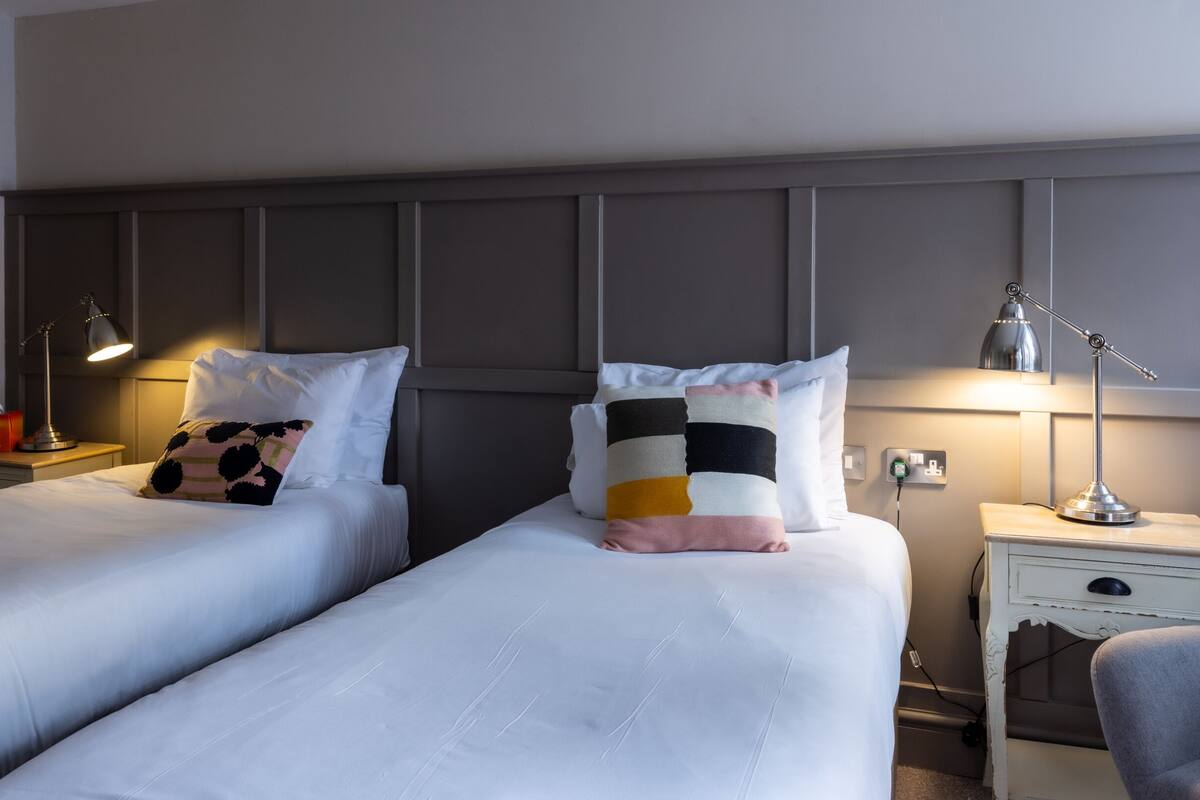 A hotel room with two beds, bedside lamps, a desk with a chair, and a power outlet.