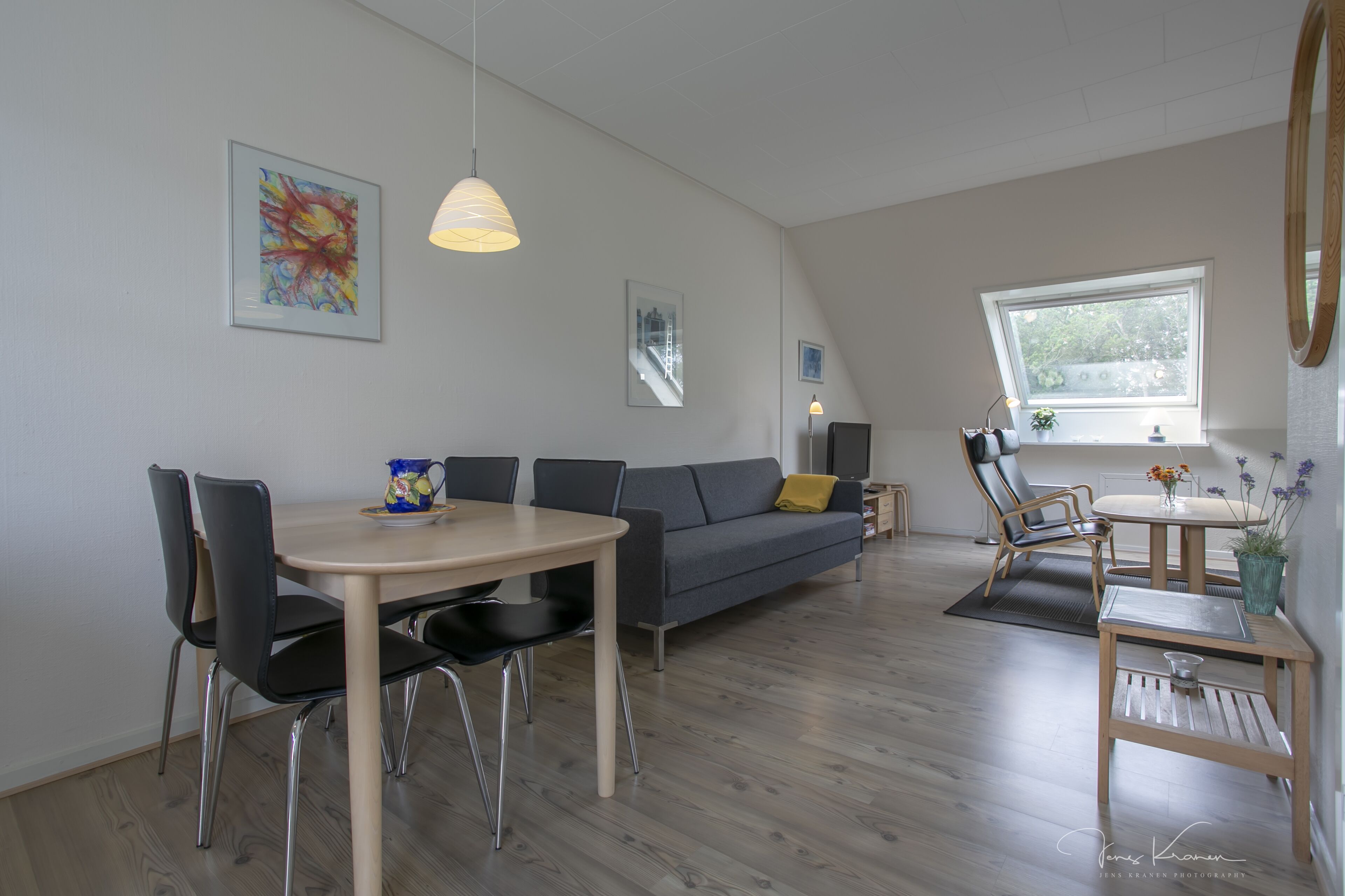 A modern living room with a dining table, sofa, and chairs. There is a window with a view of greenery outside.