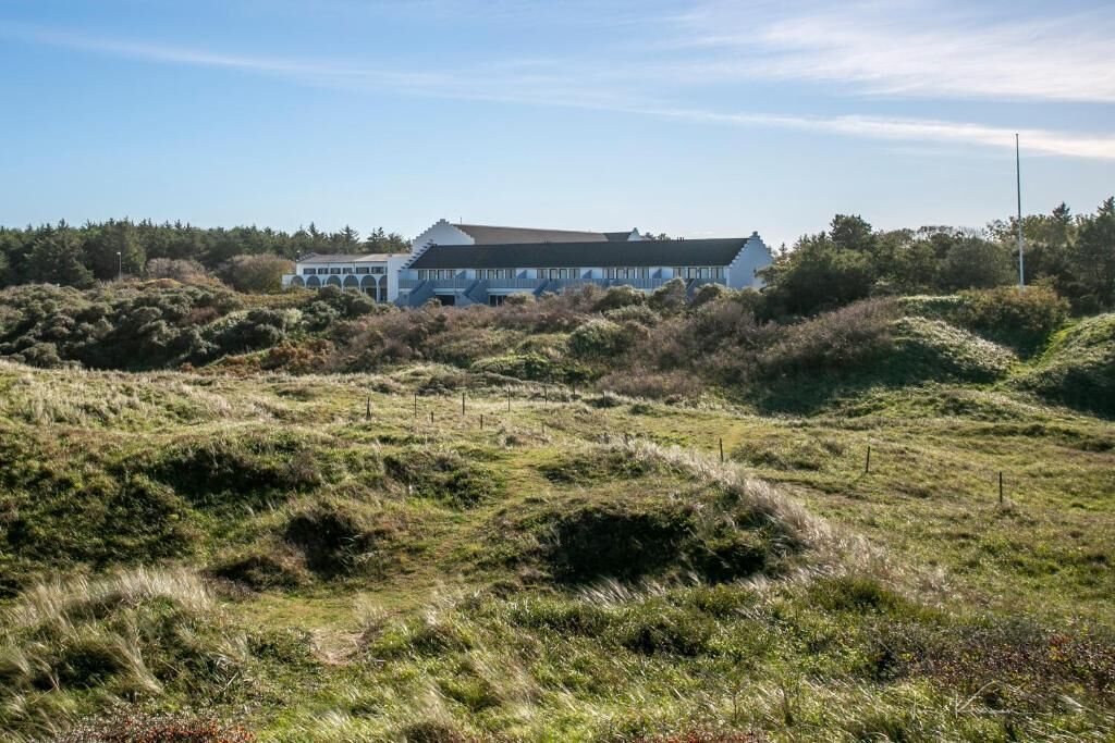 A building with a flat roof and multiple windows, surrounded by a natural landscape with undulating terrain and sparse vegetation.