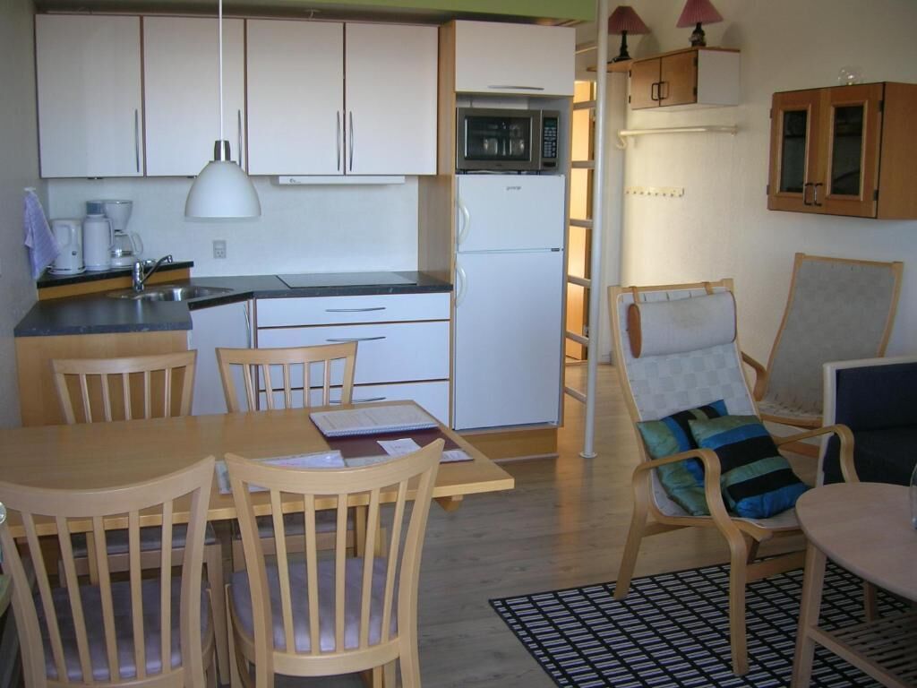 A compact kitchen with white cabinets, a microwave, and a refrigerator. There is a dining table with chairs and a patterned rug.