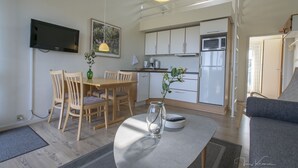 Una cocina moderna con armarios blancos, un microondas y un refrigerador. Hay un comedor con una mesa y sillas, y un televisor montado en la pared.