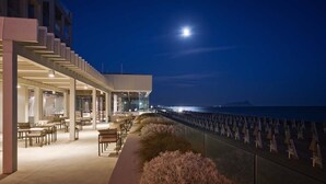 Un área de estar moderna al aire libre con mesas y sillas, con vistas a una masa de agua y una montaña lejana bajo un cielo nocturno con luna visible.