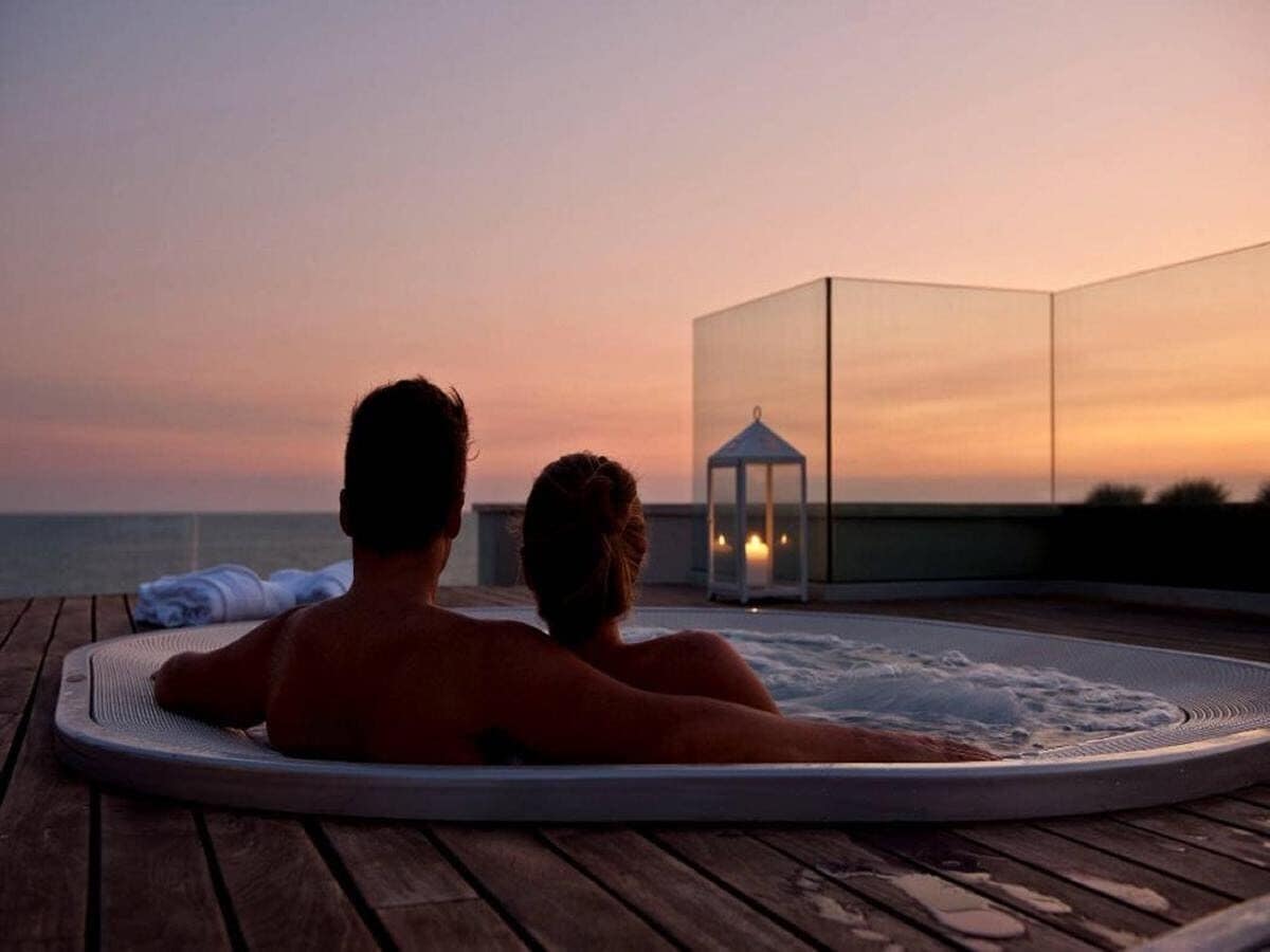 A couple relaxing in a hot tub on a wooden deck with a view of the ocean and a gazebo in the background.