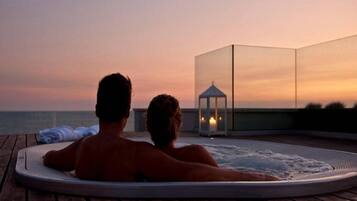 A couple relaxing in a hot tub on a wooden deck with a view of the ocean and a gazebo in the background.