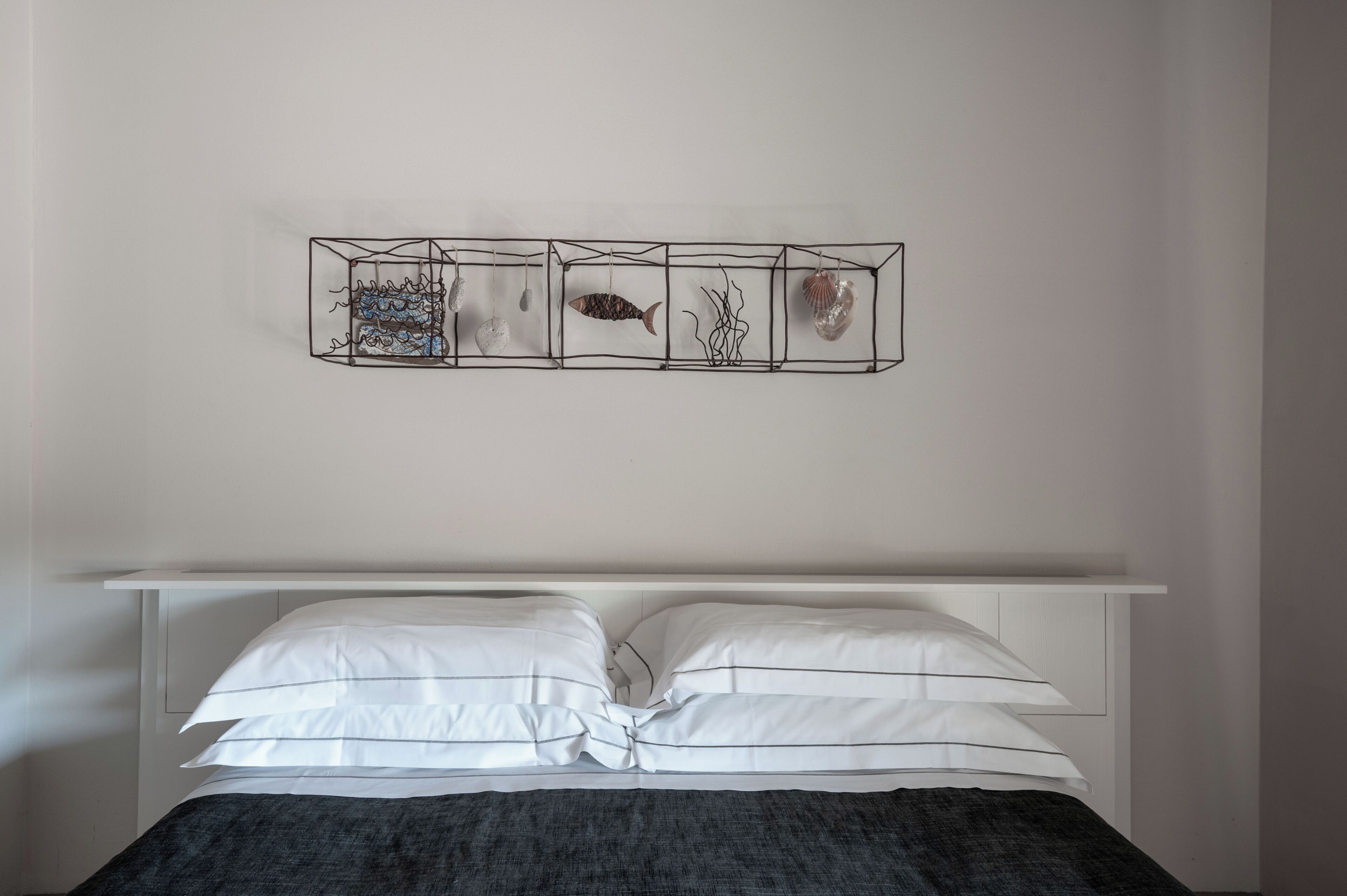 A bed with white linens and a dark blanket, a white headboard, and a wall-mounted art piece with geometric shapes and objects.