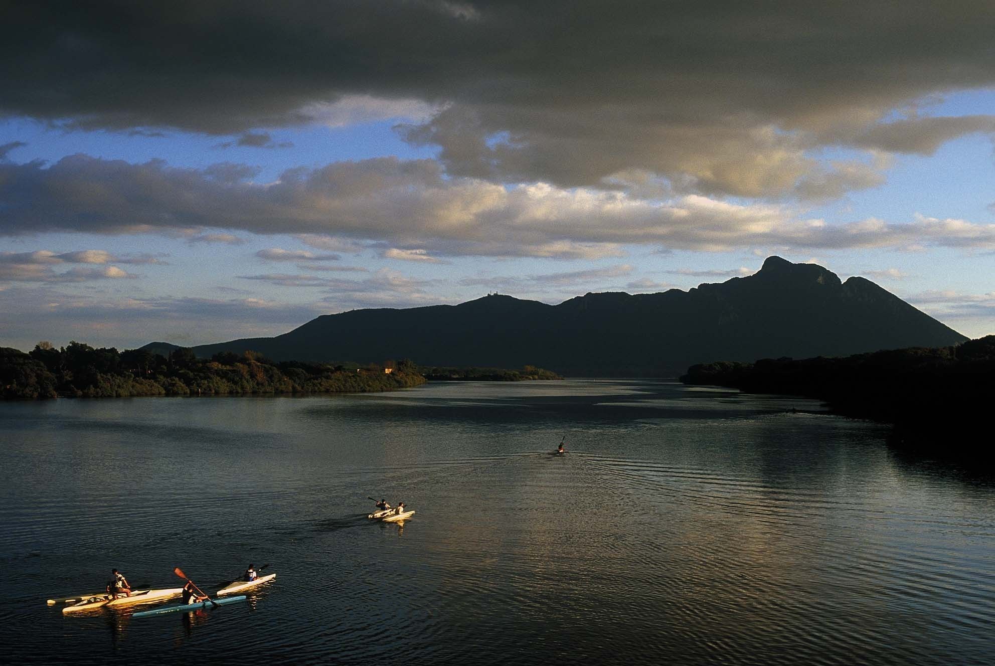 A calm lake with boats, surrounded by mountains and trees under a cloudy sky.