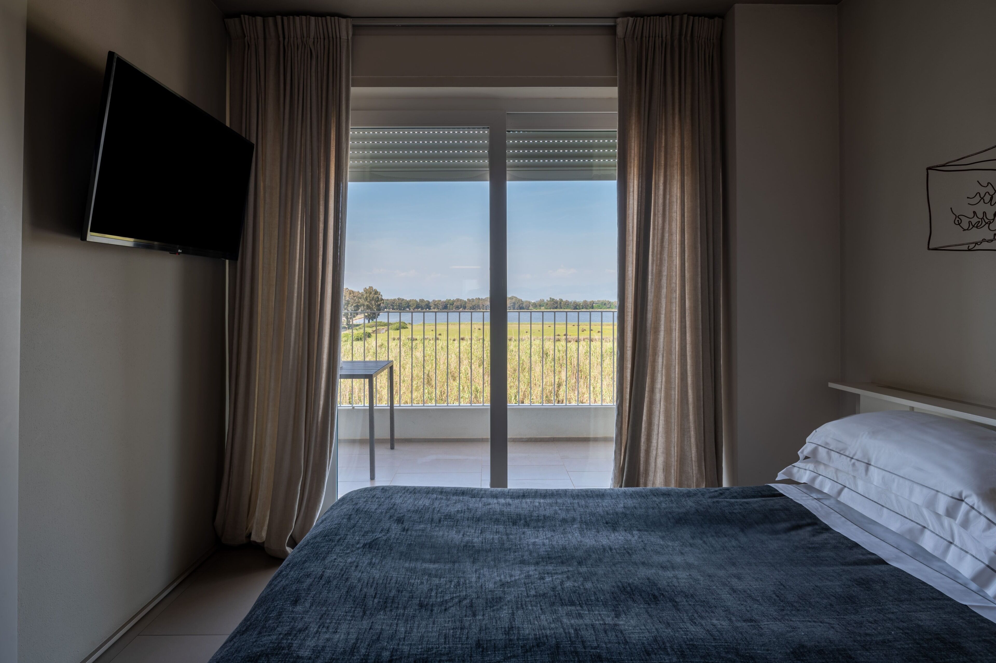 A bedroom with a bed, a TV, a balcony with curtains, and a view of a field.