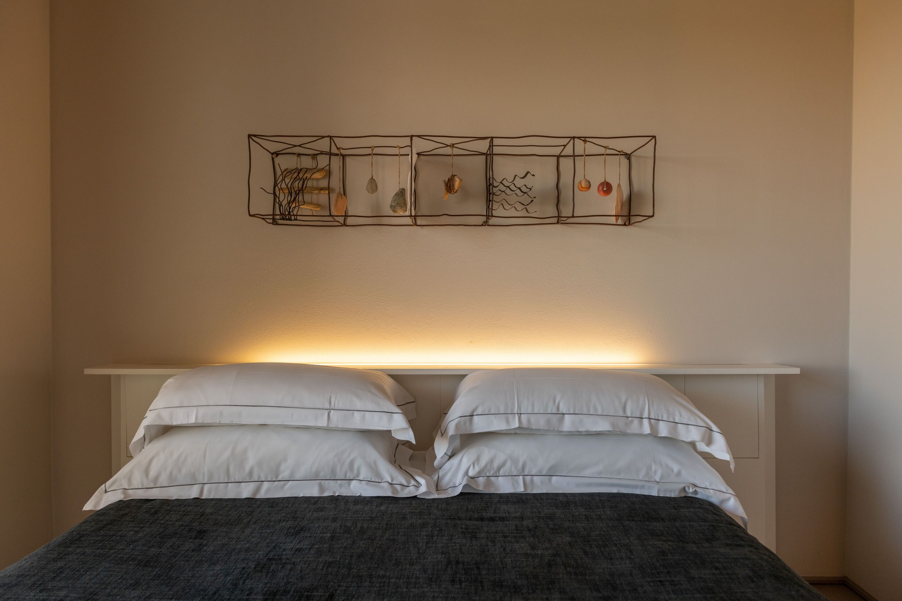 Una cama con almohadas blancas y una manta oscura, una mesita de noche y una lámpara fijada a la pared.