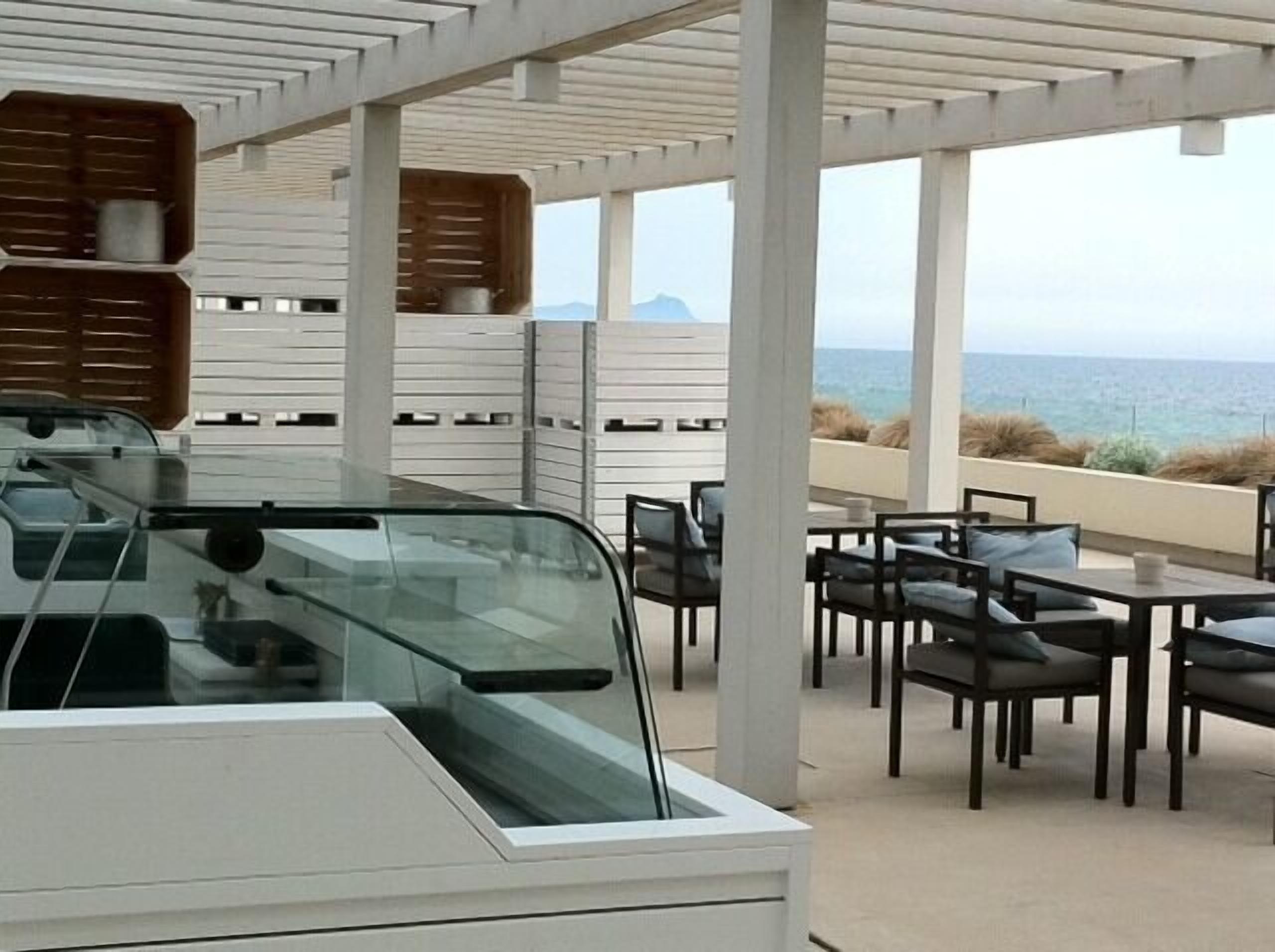 Een overdekt terras met tafels en stoelen, met uitzicht op een kustlandschap.
