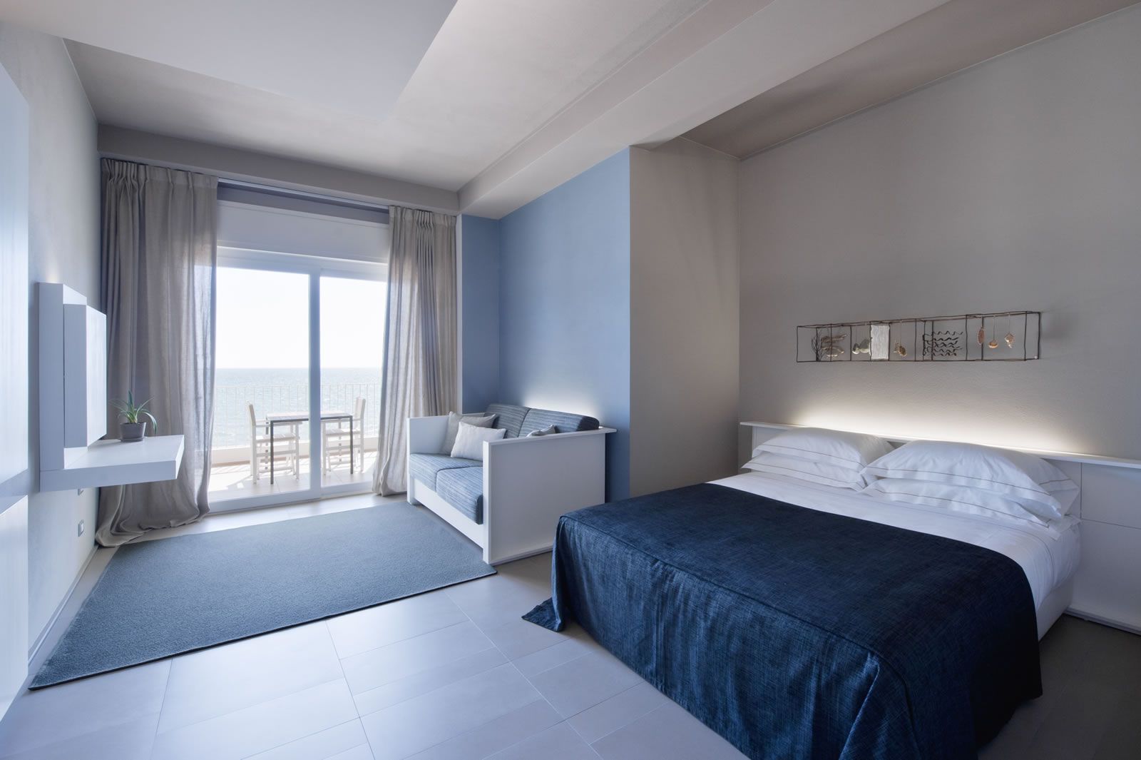 A modern hotel room with a large bed, a sofa, a balcony with a view, and a small table with a plant.