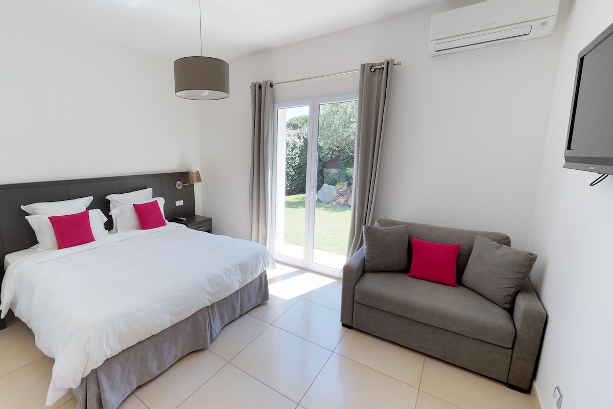 A modern hotel room with a large bed, a sofa, a nightstand, a lamp, and a view of the outdoors.
