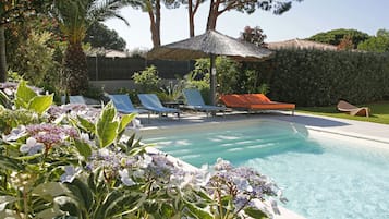 Un área de piscina con sillones de descanso, una palmera y un toldo de paja.