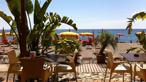 Un café frente a la playa con terraza, mesas y sillas, con vistas al mar.