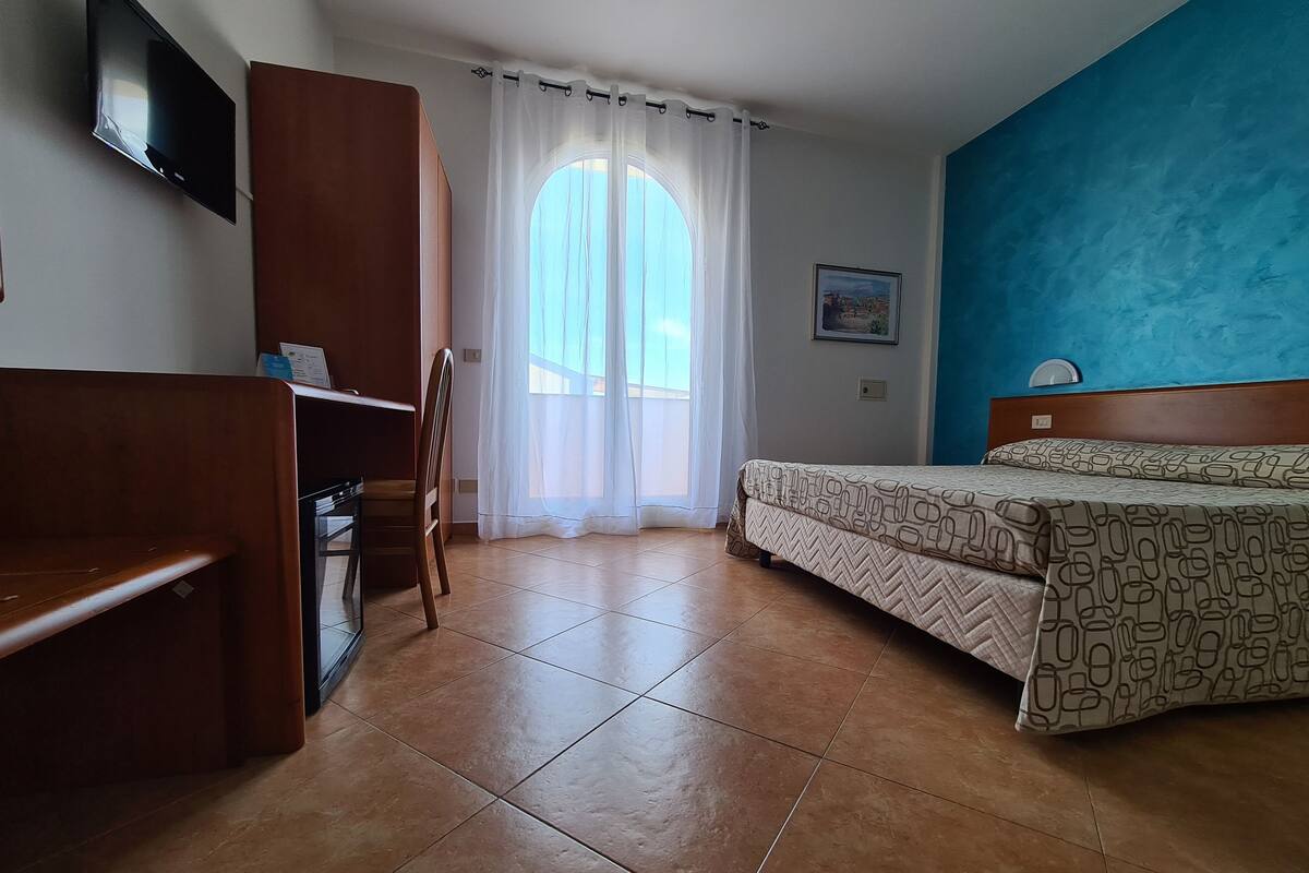 A bedroom with a bed, a desk, a chair, a TV, and a window with curtains.
